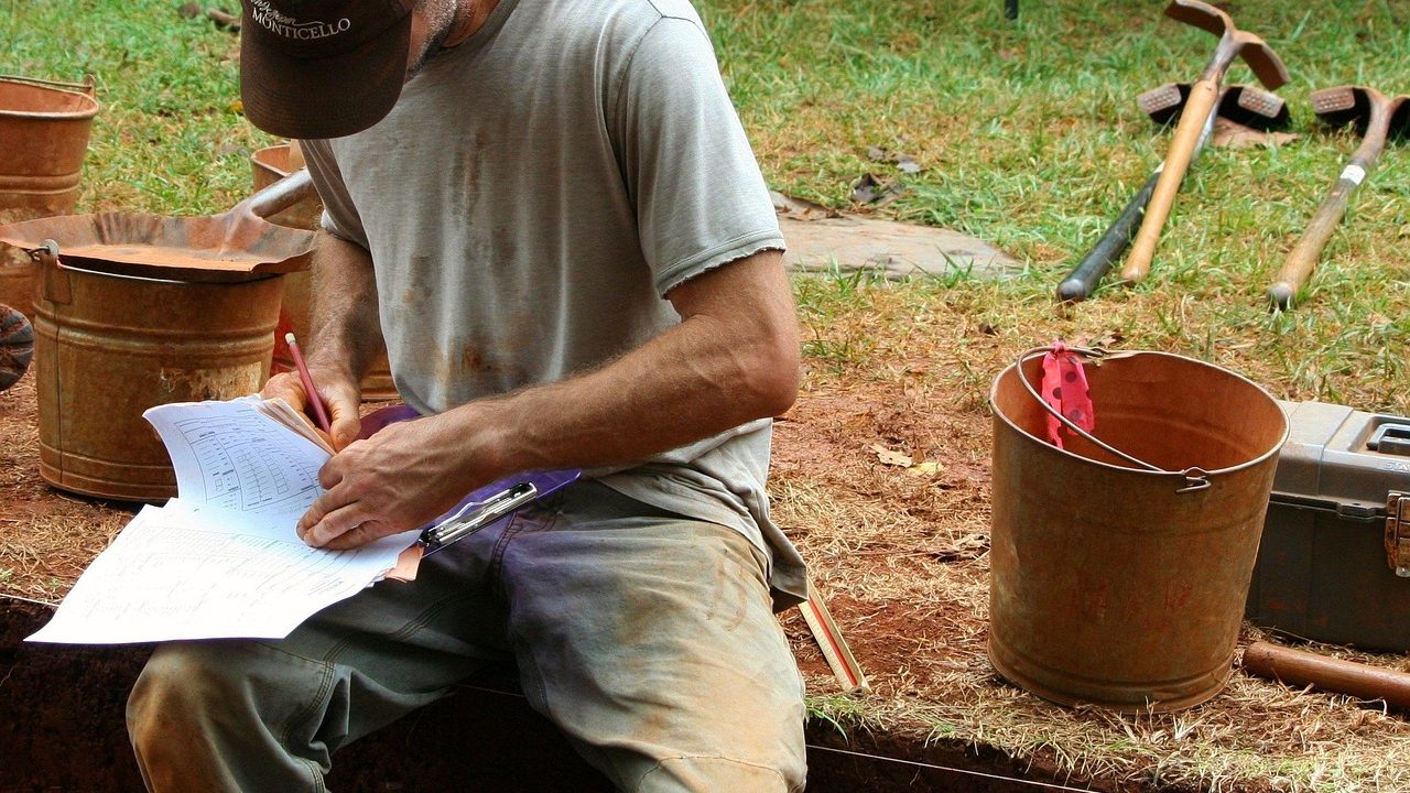 Researchers in wide-brimmed hats examining exposed stone foundations at a desert dig site.