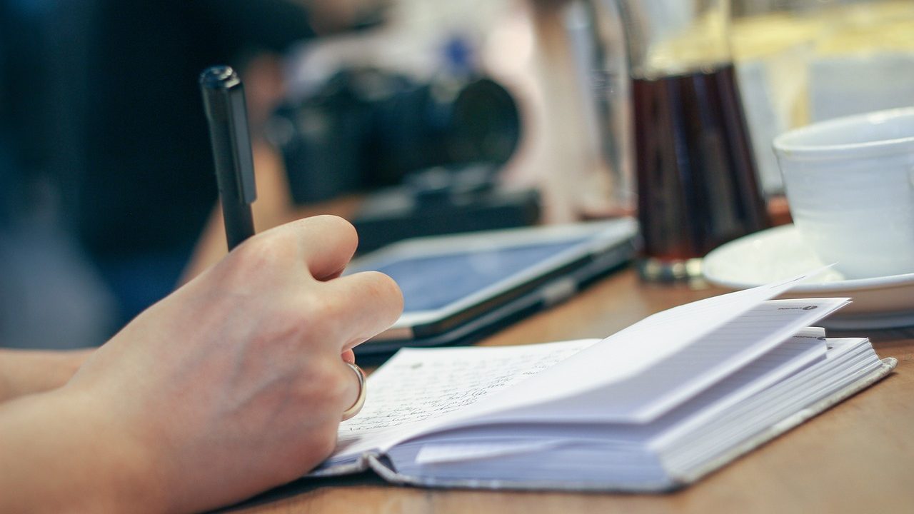 An open spiral notebook on a wooden desk next to a pen and a cup of coffee.