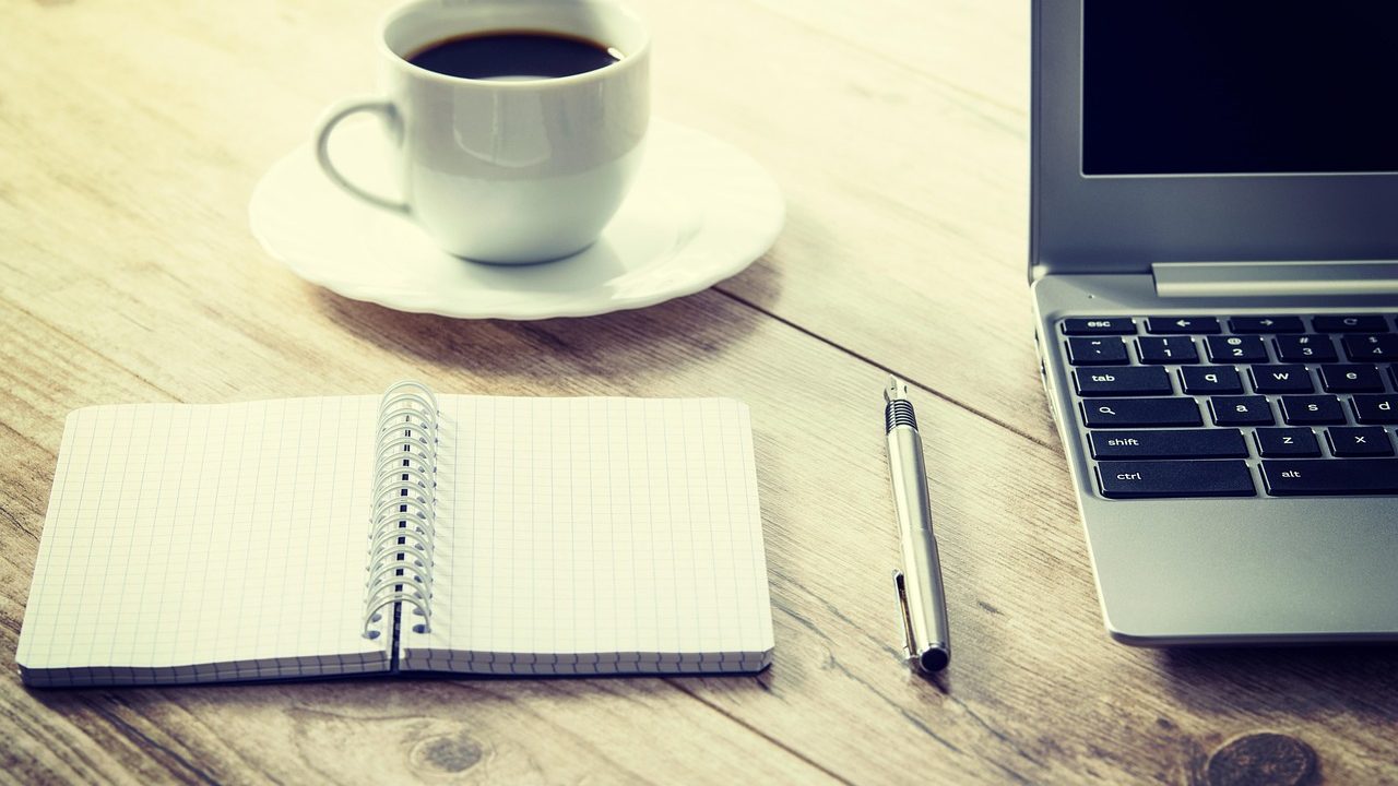 A laptop and coffee cup on a wooden desk, suggesting a morning reading session.