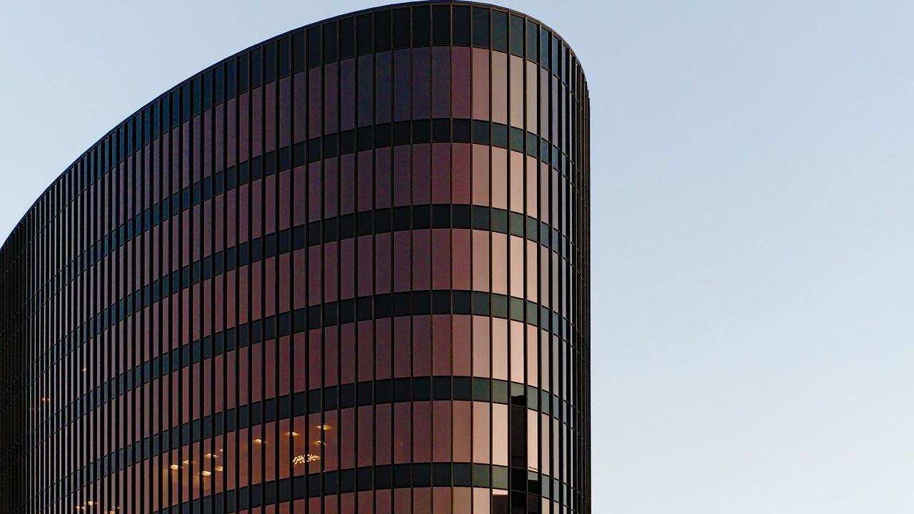 A modern glass office tower reflects blue sky, viewed from street level.