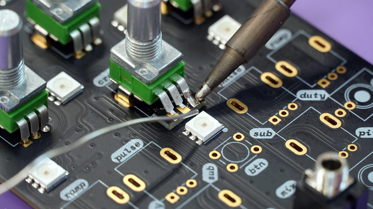 A cluttered workbench with soldering equipment, circuit boards, and electronic components.