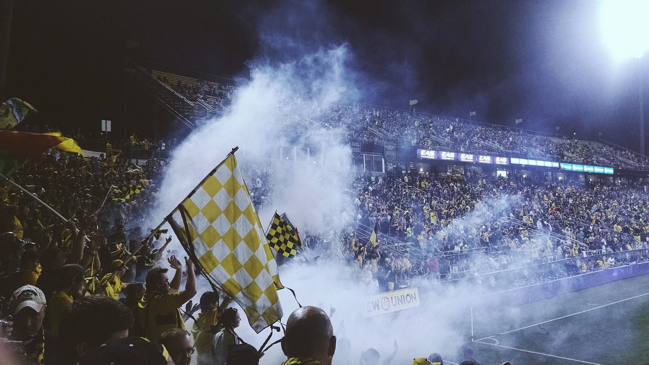 A packed soccer stadium filled with colorful flags and cheering fans under bright floodlights.