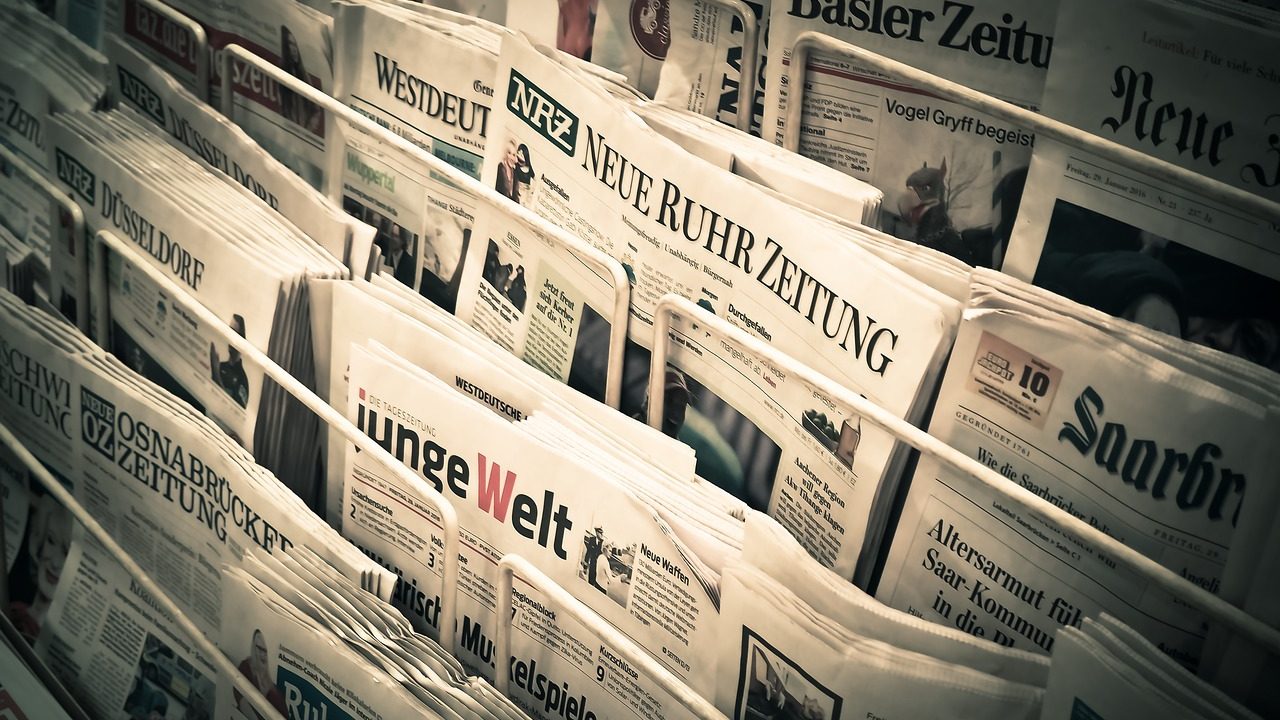 A stack of morning newspapers spread open on a table with headlines visible.