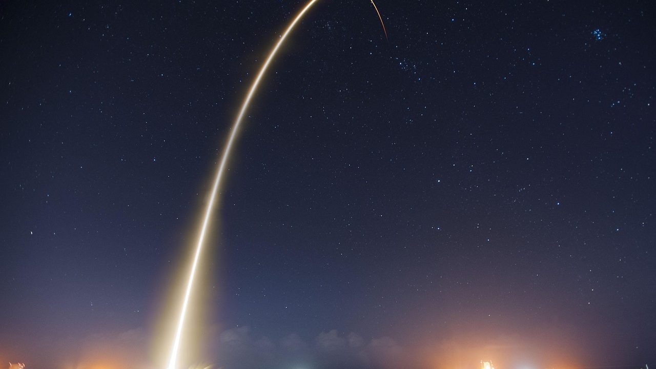 A large rocket lifts off from a launch pad, illuminating the night sky with bright exhaust flame.
