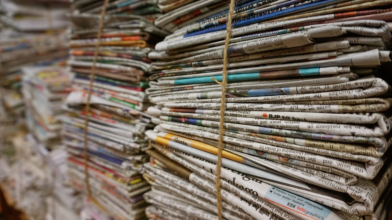 A stack of newspapers with bold headlines visible on a wooden surface.