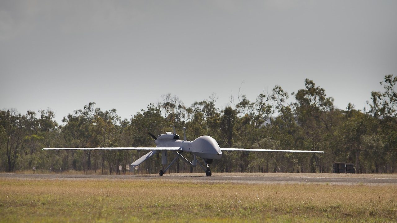 A small military-style drone flies against an overcast sky.