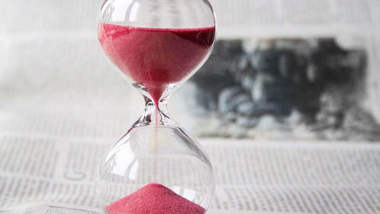 Sand grains falling through the narrow waist of a glass hourglass against a dark background.