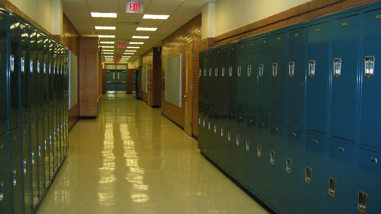 A long empty school hallway lined with closed lockers under fluorescent lighting.