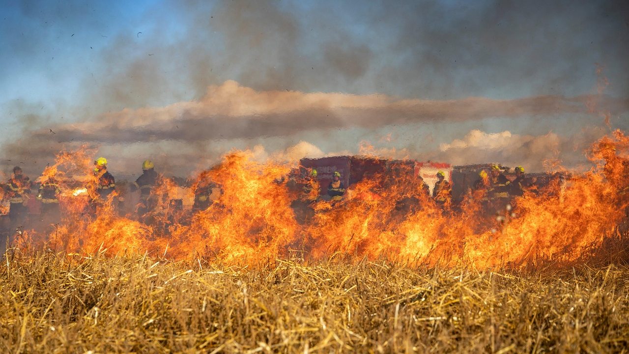 Thick brown smoke rises from a dry hillside covered in brush during an active wildfire.