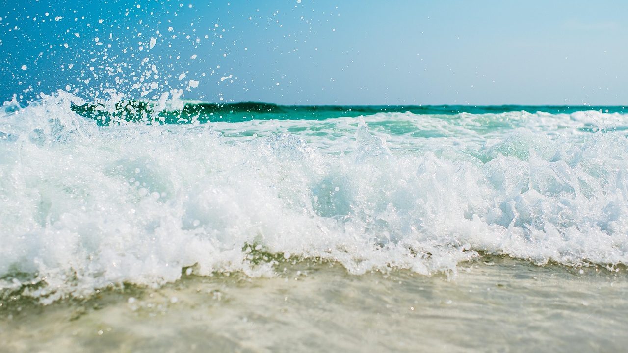 Large waves crash against a sandy beach under a partly cloudy summer sky.