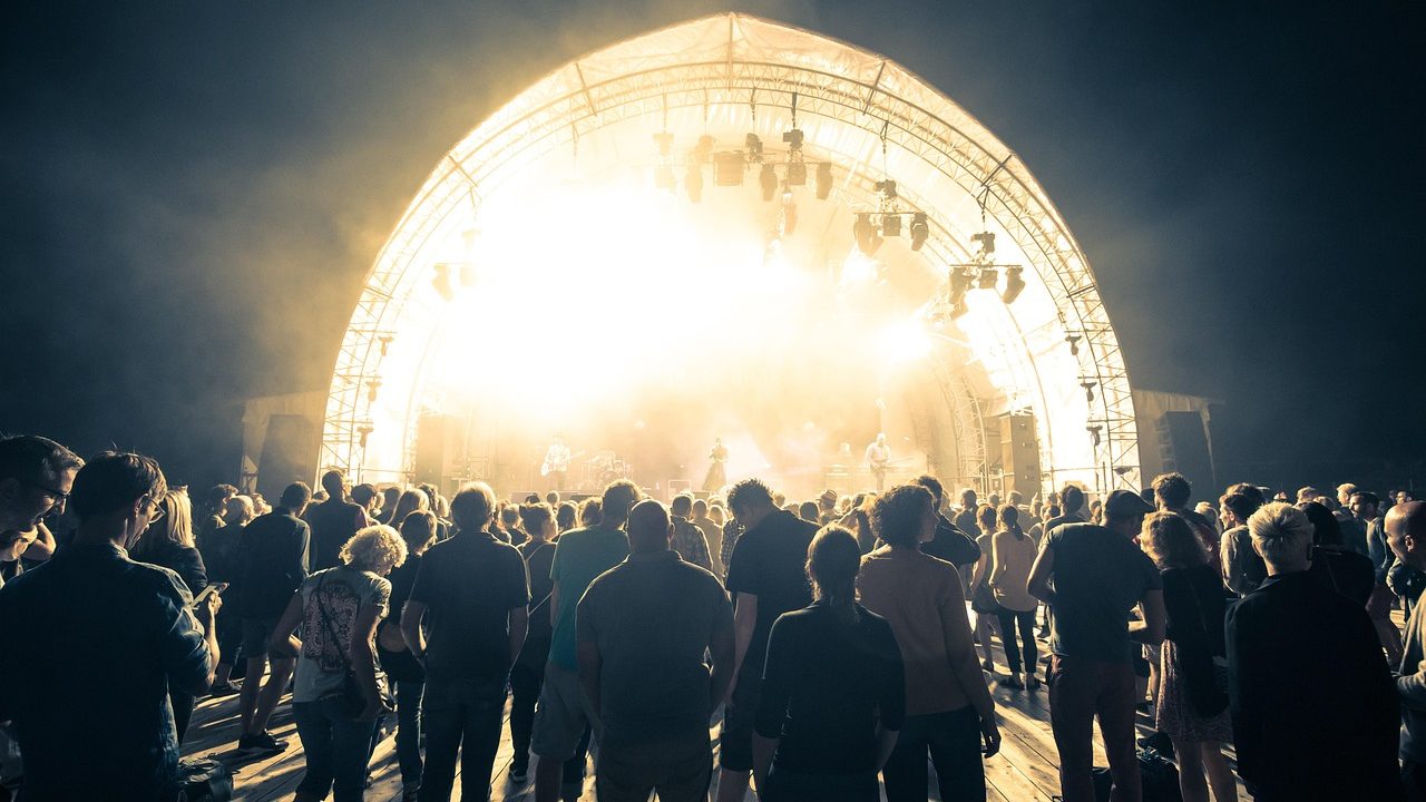 A crowd of people enjoying an outdoor concert on a warm summer evening under string lights.