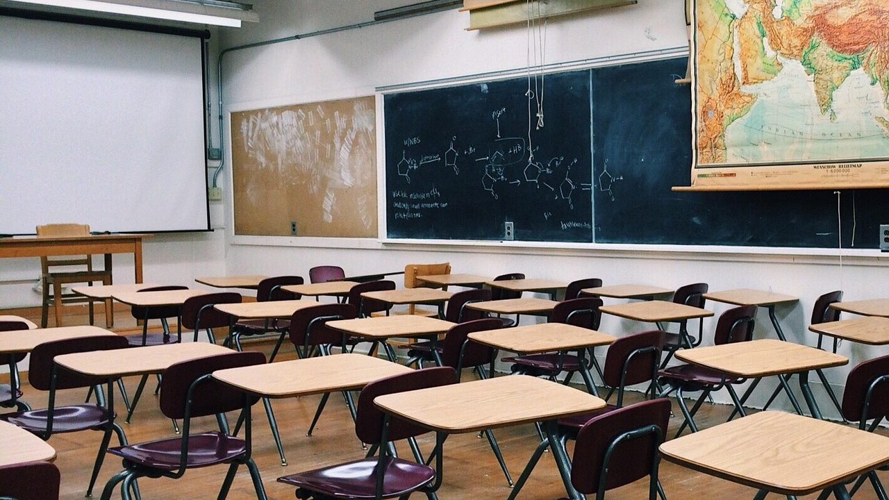 Rows of empty student desks in a sunlit elementary school classroom.