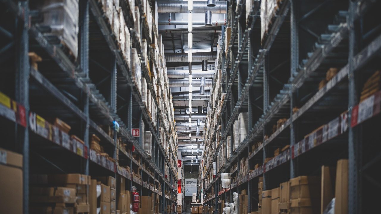 The interior of a large industrial warehouse with high ceilings, concrete floors, and stacked goods.