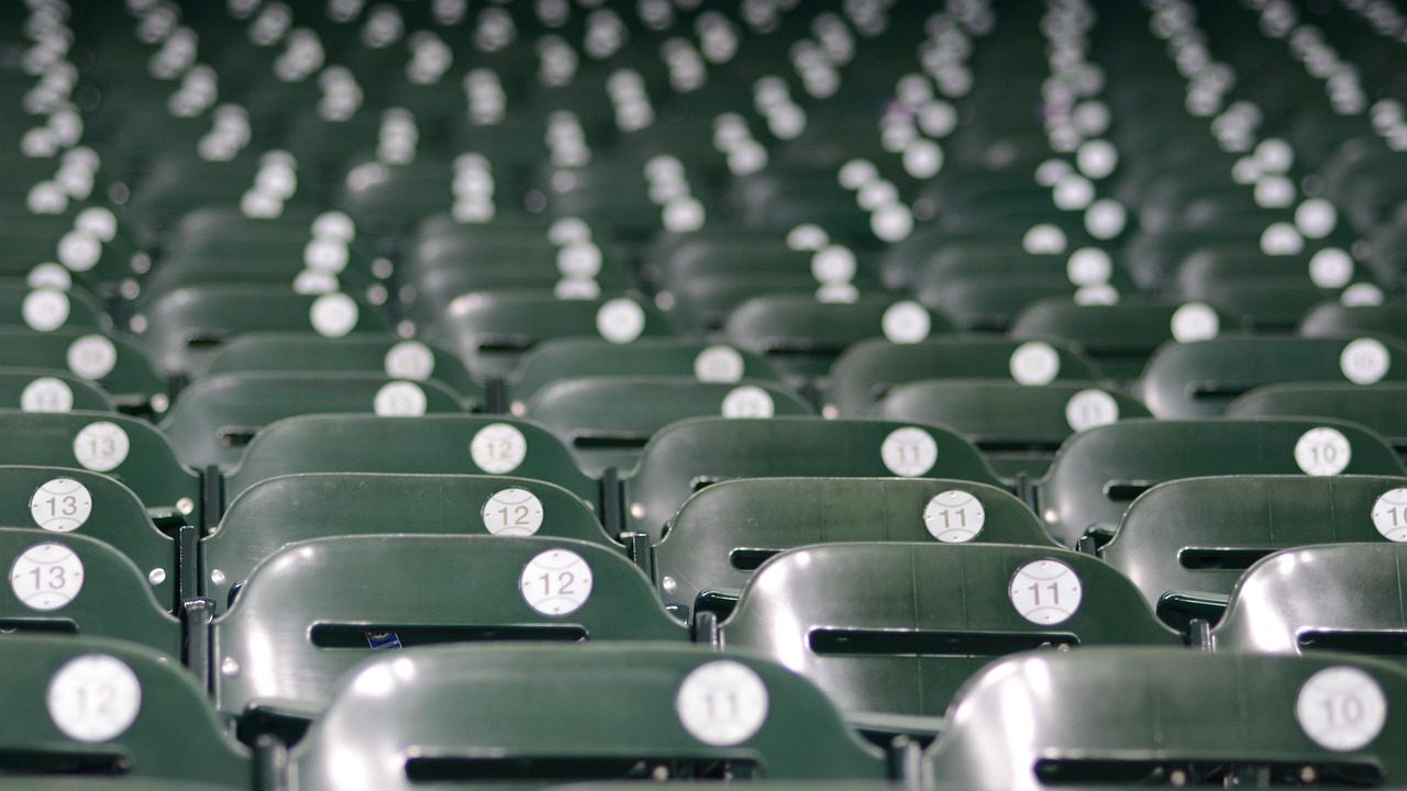 Rows of blue and orange seats fill a sunlit baseball stadium before game time.